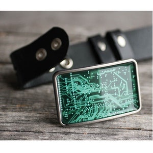 Op de afbeelding: Een rechthoekige gesp met een groen en wit printplaatontwerp, gezet in een zilverkleurig frame. De gesp wordt gecombineerd met een zwarte leren riem, een technisch ge&iuml;nspireerd accessoire.
