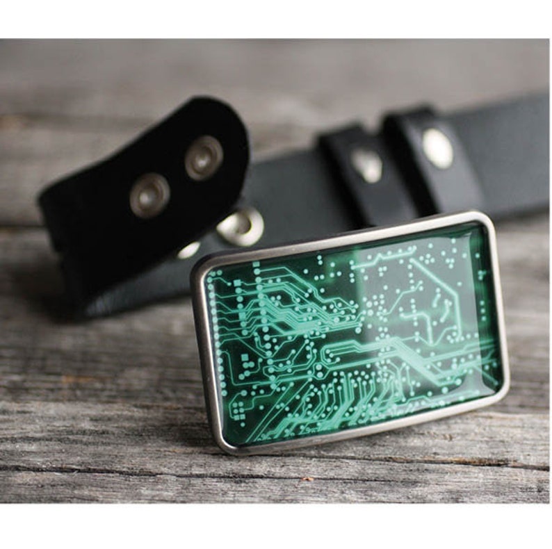 Op de afbeelding: Een rechthoekige gesp met een groen en wit printplaatontwerp, gezet in een zilverkleurig frame. De gesp wordt gecombineerd met een zwarte leren riem, een technisch ge&iuml;nspireerd accessoire.