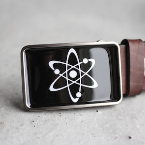 Könnte beinhalten: Eine schwarze Gürtelschnalle mit silbernem Rand und einem weißen Atom-Symbol.