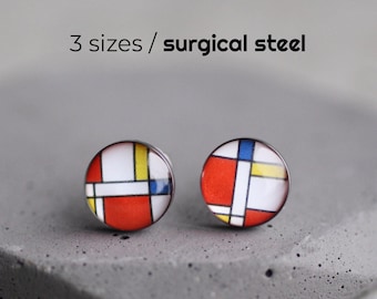 May include: A pair of round stud earrings featuring a colourful geometric design in red, white, yellow, and blue. The earrings are displayed on a grey surface. The text "3 sizes / surgical steel" is above the earrings.