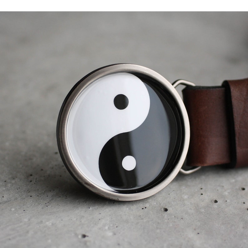 May include: A silver belt buckle with a black and white yin yang symbol. The buckle is attached to a brown leather belt.