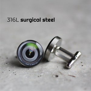 May include: A pair of silver cufflinks with a camera lens design. The lens is black with a green and white pattern. The cufflinks are made of 316L surgical steel.