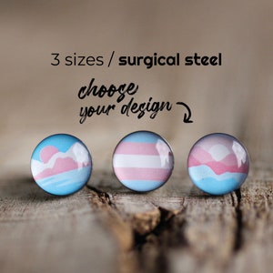May include: Three round stud earrings with various designs, including a transgender pride flag and a landscape scene in pink and blue. The text "3 sizes / surgical steel" and "choose your design" are above the earrings.