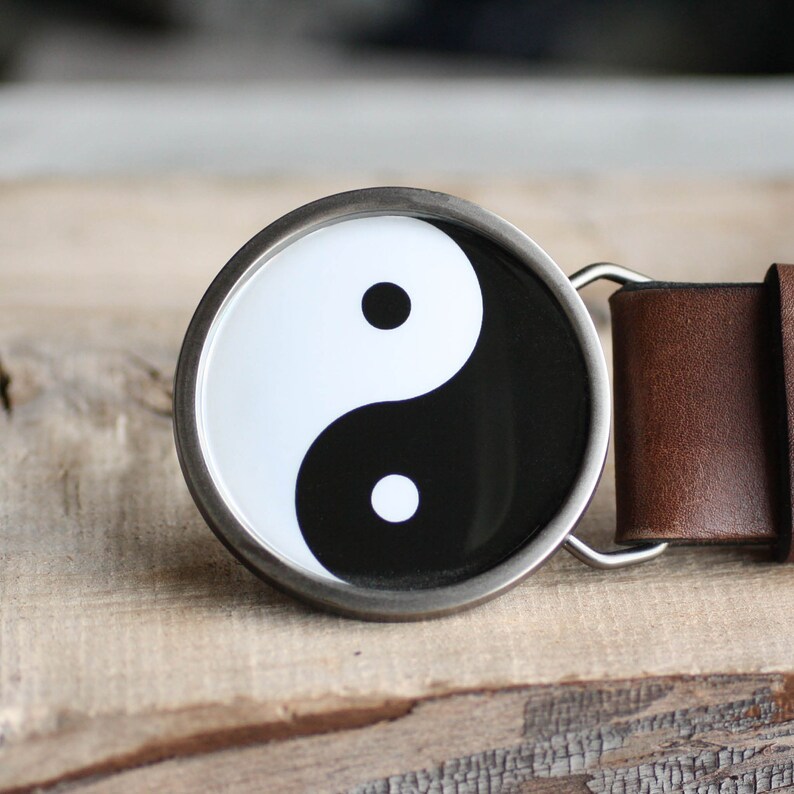 May include: A silver belt buckle with a black and white yin yang symbol. The buckle is attached to a brown leather belt.