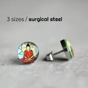 May include: Round stud earrings featuring a seated figure in a red robe, set against a stained-glass window design. The earrings are made of surgical steel. Text on the image reads: "3 sizes / surgical steel."