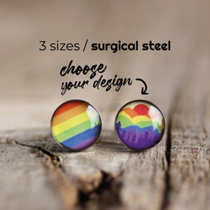 Puede incluir: Dos pendientes redondos de cristal arcoíris con postes de acero quirúrgico. Los pendientes están disponibles en tres tamaños. El pendiente izquierdo presenta un diseño de bandera arcoíris. El pendiente derecho presenta un diseño de bandera arcoíris con una cordillera y un sol en el fondo. El texto "elige tu diseño" está escrito sobre los pendientes.