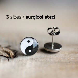 May include: A pair of yin yang earrings made of surgical steel. The earrings are black and white with a round shape. The earrings are on a wooden surface.