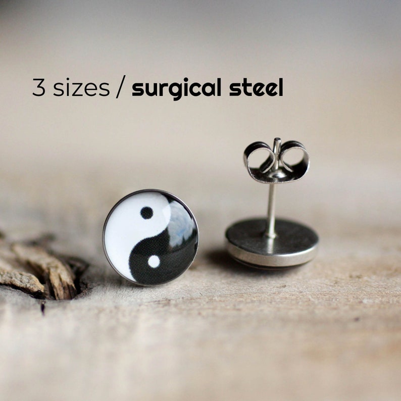May include: A pair of yin yang earrings made of surgical steel. The earrings are black and white with a round shape. The earrings are on a wooden surface.
