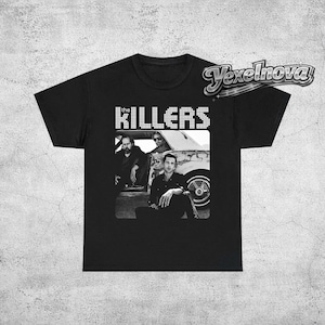 Op de afbeelding: Zwart T-shirt met een zwart-wit afbeelding van de band The Killers. De bandnaam "KILLERS" is in witte blokletters boven de afbeelding gedrukt. De afbeelding toont de bandleden die poseren voor een auto.