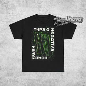 May include: Black t-shirt featuring a green graphic of two coffins with a skeleton and the text "TYPE O NEGATIVE" and "DEAD AGAIN" in white. The design has a grunge aesthetic.