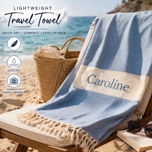 Könnte beinhalten: Ein blau-weißes, leichtes Reisehandtuch mit dem gestickten Namen "Caroline", das über einem Strandstuhl drapiert ist. Das Handtuch hat ein Rautenmuster und Fransen. Eine Strohtasche, Sonnenbrille und ein Buch liegen ebenfalls auf dem Stuhl.