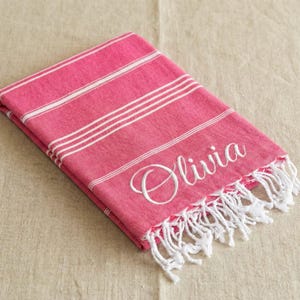 Peut inclure: Serviette de plage rose pliée avec des rayures blanches et des franges. Le nom "Olivia" est brodé en écriture blanche. La serviette est en matière douce et est pliée avec soin.