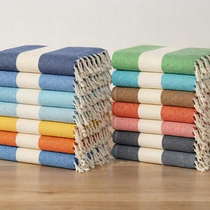 Könnte beinhalten: Zwei Stapel türkischer Handtücher mit Streifenmustern. Ein Stapel hat blaue, hellblaue, türkisfarbene, gelbe und orangefarbene Streifen. Der andere hat grüne, türkisfarbene, braune, rote, rosa und graue Streifen. Jedes Handtuch hat einen weißen Streifen und Fransen.