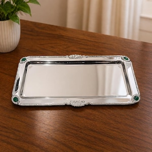 Könnte beinhalten: Ein rechteckiges Serviertablett aus Silber mit dekorativen Kanten und grünen Akzenten. Das Tablett hat eine reflektierende Oberfläche und steht auf einem dunklen Holztisch. Die Ecken weisen kleine, runde grüne Verzierungen auf.