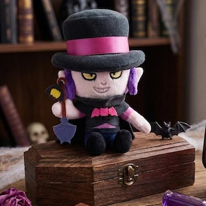 Puede incluir: Un muñeco de peluche con sombrero de copa negro con banda rosa, capa negra con emblema de murciélago rosa y ropa oscura. El muñeco sostiene una pequeña pala azul. A su lado hay un farol de color bronce con un inserto de vidrio de color púrpura.