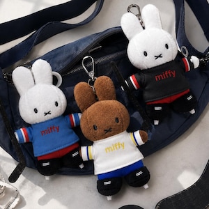 May include: Three plush Miffy rabbit keychains with metal clips attached to a navy blue crossbody bag. One white rabbit wears a blue sweater, one brown rabbit wears a white sweater, and one white rabbit wears a black sweater, all with the text miffy.