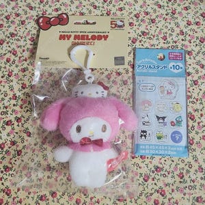 May include: A My Melody plush keychain with a white Hello Kitty figure on its head, featuring a pink hood, a red bow tie, and a white body. The plush is in a clear plastic bag with a Hello Kitty 50th Anniversary header card and a separate acrylic stand pack.