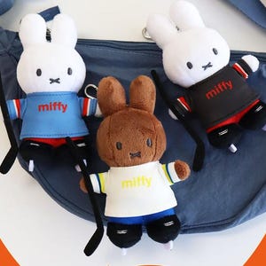 Puede incluir: Tres juguetes de peluche con forma de conejito, cada uno con un palo de hockey. Dos conejos blancos visten camisetas azules y negras con "miffy" impreso. El tercer conejo es marrón y lleva una camiseta blanca con "miffy" impreso. Los tres tienen patines negros.