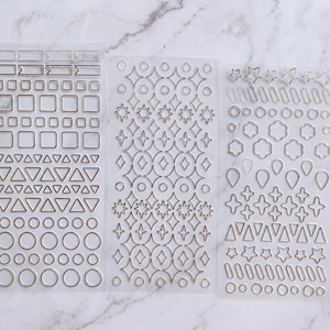 Könnte beinhalten: Drei transparente Aufkleberbögen mit metallisch silbernen Umrissen verschiedener geometrischer Formen. Die Designs umfassen Rechtecke, Quadrate, Dreiecke, Kreise, Sterne, vierzackige Funken, Blumenumrisse und tropfenförmige Elemente in Reihen.