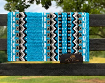 Op de afbeelding: Een turquoise en patroon zadeldek met een zwart, wit en grijs ontwerp. Het dek wordt getoond op een zwarte hek met een leren label met de tekst "THE EQUI STORE". Het dek is ontworpen voor paardensport.