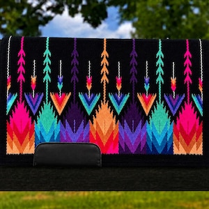 Könnte beinhalten: Eine rechteckige schwarze Satteldecke mit einem lebendigen, mehrfarbigen geometrischen Muster aus Pfeilen und Dreiecksformen in Neonrosa, Türkis, Violett und Orange sowie einem einfarbigen schwarzen, lederartigen Aufnäher unten in der Mitte.