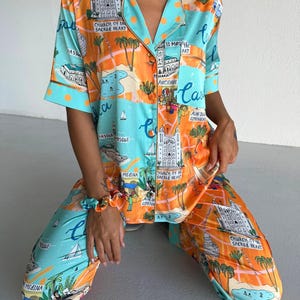 May include: A two-piece pajama set in a vibrant orange and turquoise color scheme, featuring a map-like design with architectural illustrations and text. The set includes a button-down shirt and matching pants. A scrunchie in matching colors is also visible.