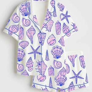 Puede incluir: Conjunto de pijama blanco con un estampado de conchas marinas en rosa y azul. El conjunto incluye una camisa de manga corta con botones y pantalones cortos a juego. Los bordes están ribeteados con ribete azul.