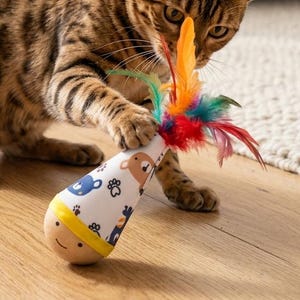Puede incluir: Un gato bengalí marrón y negro jugando con un juguete para gatos. El juguete es blanco con ilustraciones de osos azules y plumas de colores. El gato está sobre un suelo de madera.