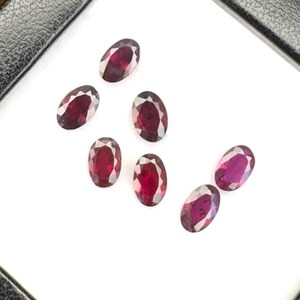May include: Six oval-cut, deep red gemstones are arranged on a white display insert within a black, square-shaped storage case. The faceted stones exhibit a rich, dark crimson hue and are positioned centrally against the white background.