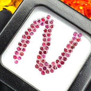 May include: A collection of small, round, faceted deep pink gemstones arranged in a winding, serpentine pattern inside a square black display case with a white interior base. The stones exhibit a consistent vibrant magenta hue.