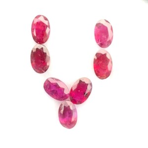 May include: Six small, oval-cut, vibrant pinkish-red gemstones arranged in a V-shape inside a square black display case with a white interior. The faceted stones exhibit a polished, reflective surface and are positioned centrally within the frame.