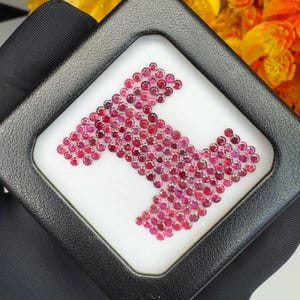 Può includere: Una collezione di piccole pietre preziose rosse, rotonde e sfaccettate, disposte a forma di lettera maiuscola H. Le pietre sono esposte all'interno di una custodia quadrata nera con rivestimento interno bianco, che ne esalta il colore cremisi.
