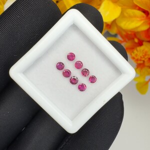 Op de afbeelding: Een verzameling van zeven kleine, ronde, gefacetteerde magentakleurige edelstenen gerangschikt in een vierkante witte displaydoos. De edelstenen vertonen een levendige roze-rode tint tegen een witte achtergrond.