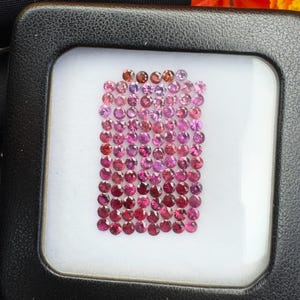 May include: A collection of small, round, faceted gemstones arranged in a rectangular grid inside a black display case. The stones exhibit a gradient of colors ranging from light pink and lavender at the top to deep magenta and dark red at the bottom.