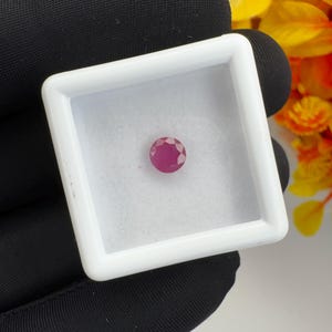 Può includere: Una piccola pietra preziosa rosa, rotonda e sfaccettata, centrata all'interno di una scatola espositiva quadrata bianca. La pietra presenta un taglio brillante con molteplici sfaccettature triangolari che riflettono la luce.