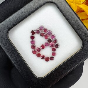 May include: A collection of small, round, faceted red gemstones arranged in a heart shape inside a square black display box with a white foam insert. The stones exhibit varying shades of deep red and pink, each featuring multiple reflective facets.