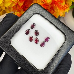 May include: A collection of six small, oval-cut, deep red gemstones arranged on a white display surface inside a square black case with a clear lid. The faceted stones exhibit a dark crimson hue and are positioned in a cluster within the box.