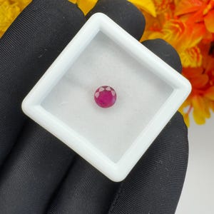 Könnte beinhalten: Ein runder, facettierter, tief pink-roter Edelstein in einer quadratischen weißen Kunststoffschatulle mit transparentem Deckel. Der Edelstein weist einen Brillantschliff mit mehreren dreieckigen Facetten auf, die das Licht reflektieren.