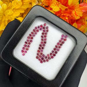 May include: A collection of small, round, faceted red gemstones arranged in a serpentine pattern inside a square black display box with a white foam insert. The stones exhibit a deep crimson hue and reflective surfaces, showcasing a uniform cut.