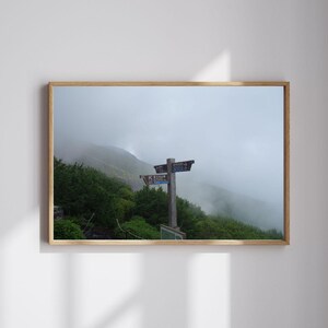以下が含まれることがあります： 木製のフレームに入った、霧のかかった山岳風景のプリント。 方向を示す標識が付いた木製の標識が前景にあり、緑の葉に囲まれています。 空は曇っています。