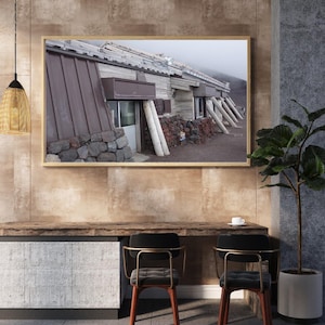 May include: A framed photograph of a rustic building with a brown roof and stone foundation. The building is supported by wooden beams and surrounded by volcanic rock. The image is displayed above a wooden desk with two chairs.