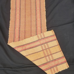 May include: A woven placemat with a scalloped edge. The placemat features vertical stripes in shades of brown, red, and tan. A portion of the placemat is folded over, revealing a plaid pattern in similar colors. The background is black.