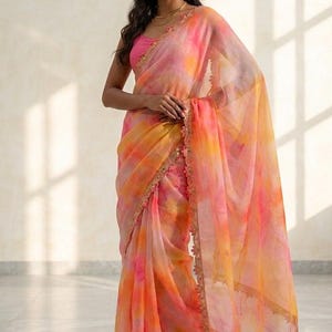 Könnte beinhalten: Ein lebendiger Batik-Sari in Rosa-, Orange- und Gelbtönen. Der transparente Stoff hat einen zarten Goldbesatz an den Kanten. Der Sari wird mit einem passenden rosa Oberteil und Goldschmuck getragen.