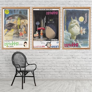 Op de afbeelding: Drie ingelijste posters met kunstwerken uit de animatiefilm "My Neighbor Totoro" hangen aan een witte bakstenen muur. De posters hebben houten frames en tonen scènes met personages en Japanse tekst. Een zwarte rieten stoel staat voor de muur.