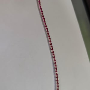 Puede incluir: Una pulsera de tenis en tono plateado que presenta una hilera continua de pequeñas piedras preciosas redondas de color rojo magenta vibrante engastadas en un marco metálico, con un cierre seguro en el extremo superior.