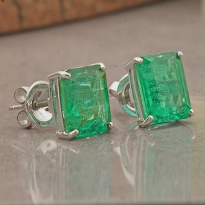 May include: A pair of emerald-cut emerald stud earrings. The bright green gemstones are set in a silver metal. The earrings have a post and butterfly backing. The earrings are displayed on a reflective surface.