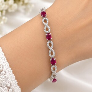 May include: A silver-toned bracelet featuring alternating oval-cut deep red gemstones and infinity-shaped links encrusted with small clear white stones. The design creates a continuous pattern of red stones and sparkling infinity symbols.