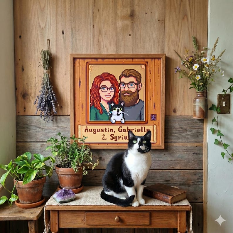 Puede incluir: Un retrato pixelado enmarcado de una pareja y un gato, con los nombres "Augustin, Gabriella & Syriu" debajo. Un gato blanco y negro est&aacute; sentado en una mesa de madera frente al retrato. La mesa tiene un caj&oacute;n y un libro peque&ntilde;o.