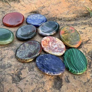 Könnte beinhalten: Eine Sammlung polierter, oval geformter Edelsteine in verschiedenen Farben, darunter Rot, Blau, Grün und gemusterte Steine. Die Steine sind auf einer rauen, braunen Oberfläche angeordnet und zeigen ihre natürlichen Texturen und Farbtöne.