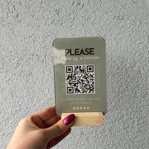Pode incluir: Uma placa retangular de acrílico verde sálvia com base de madeira, apresentando o texto preto PLEASE Leave us a review, um código QR central, o texto Scan the QR code with your phone camera e cinco estrelas douradas para avaliações.